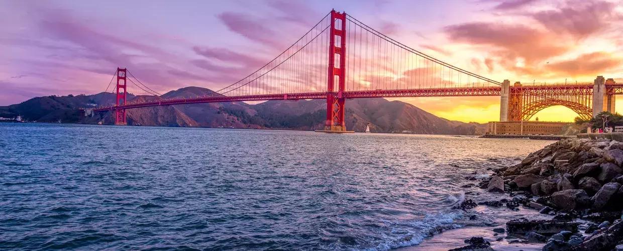 A 金门大桥 ao pôr do sol com um céu multicolorido e a Baía de São Francisco em primeiro plano.